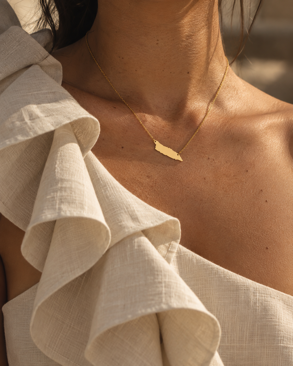 A close-up lifestyle shot of a model wearing a delicate gold necklace with a small, flat gold Aruba pendant and textured off-white ruffled top.