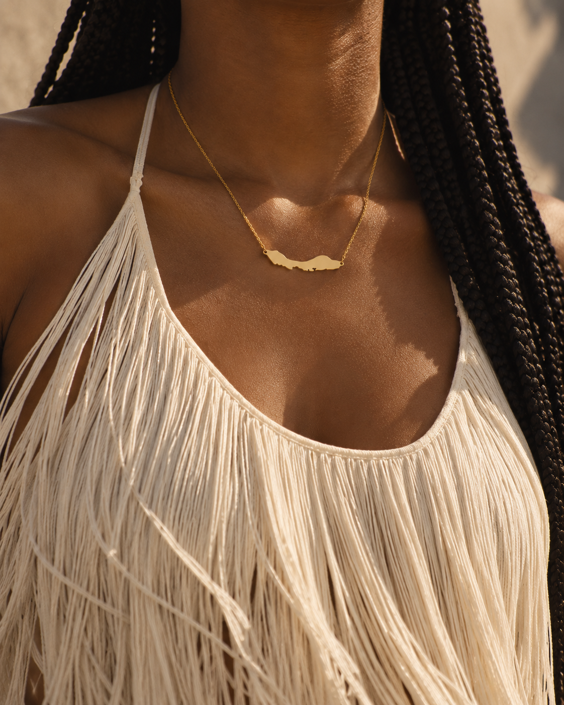 A close-up lifestyle shot of a model wearing a delicate gold necklace with a small, polished map-shaped Curacao pendant. The model is wearing a cream-colored fringed halter top, and her dark braided hair is visible/