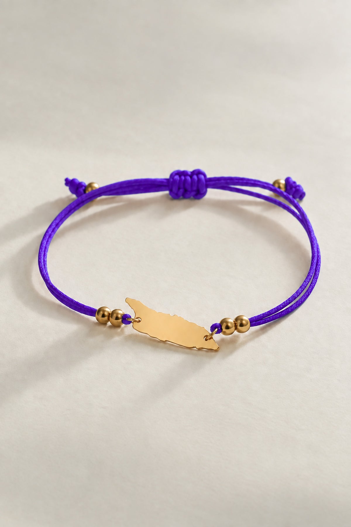 A purple cord bracelet with a small gold-tone silhouette Aruba charm and gold beads, photographed on a light neutral background.