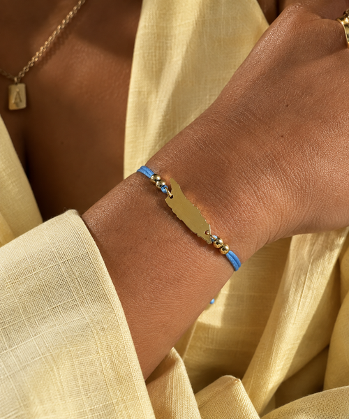 A close-up of a person's wrist wearing a gold-plated map of Aruba charm bracelet with a blue cord and small gold beads, styled with a cream linen top.