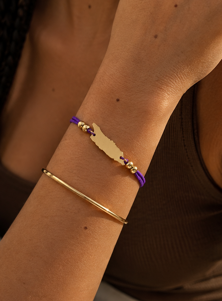 A close-up of a person's forearm wearing two bracelets: a gold-plated Aruba map-shaped charm bracelet with purple and gold beads, and a slim gold bangle, against a warm brown top and white pants.