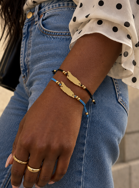 A close-up of a person's wrist wearing three bracelets: gold-plated Aruba black cords and one gold-plated Aruba on a blue cord, paired with a white polka-dot blouse and blue jeans.