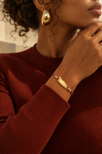 A close-up lifestyle shot of a woman wearing Aruba bracelet on her wrist, paired with a rust-red ribbed sweater. She is also wearing a gold teardrop earring.
