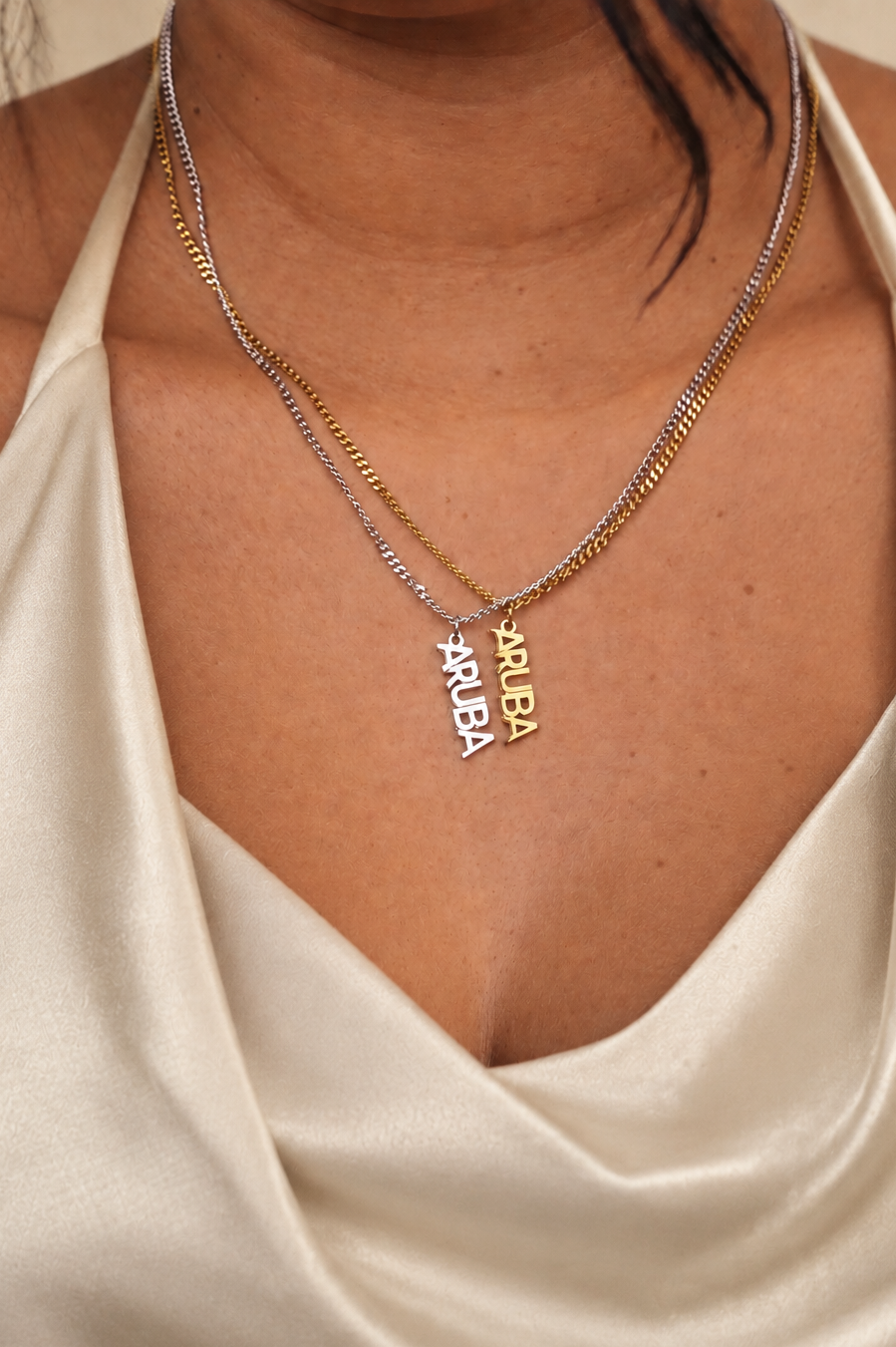 A close-up of a person wearing two delicate necklaces over a cream satin halter top. One necklace is silver-toned and the other is gold-toned, both featuring vertical letter pendants spelling 'Aruba'.