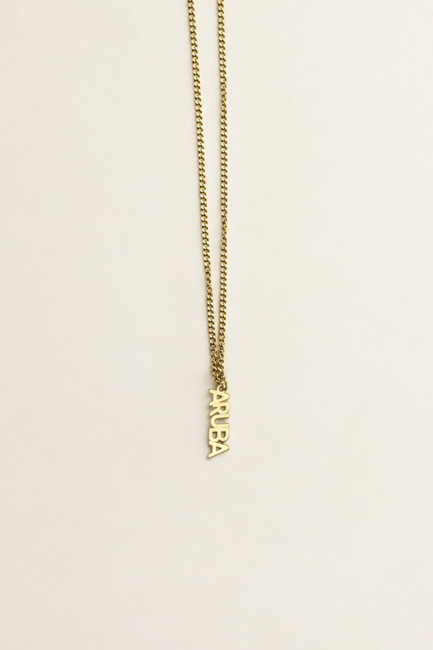 A gold chain necklace with a small vertical pendant spelling 'ARUBA' displayed against a solid off-white background.