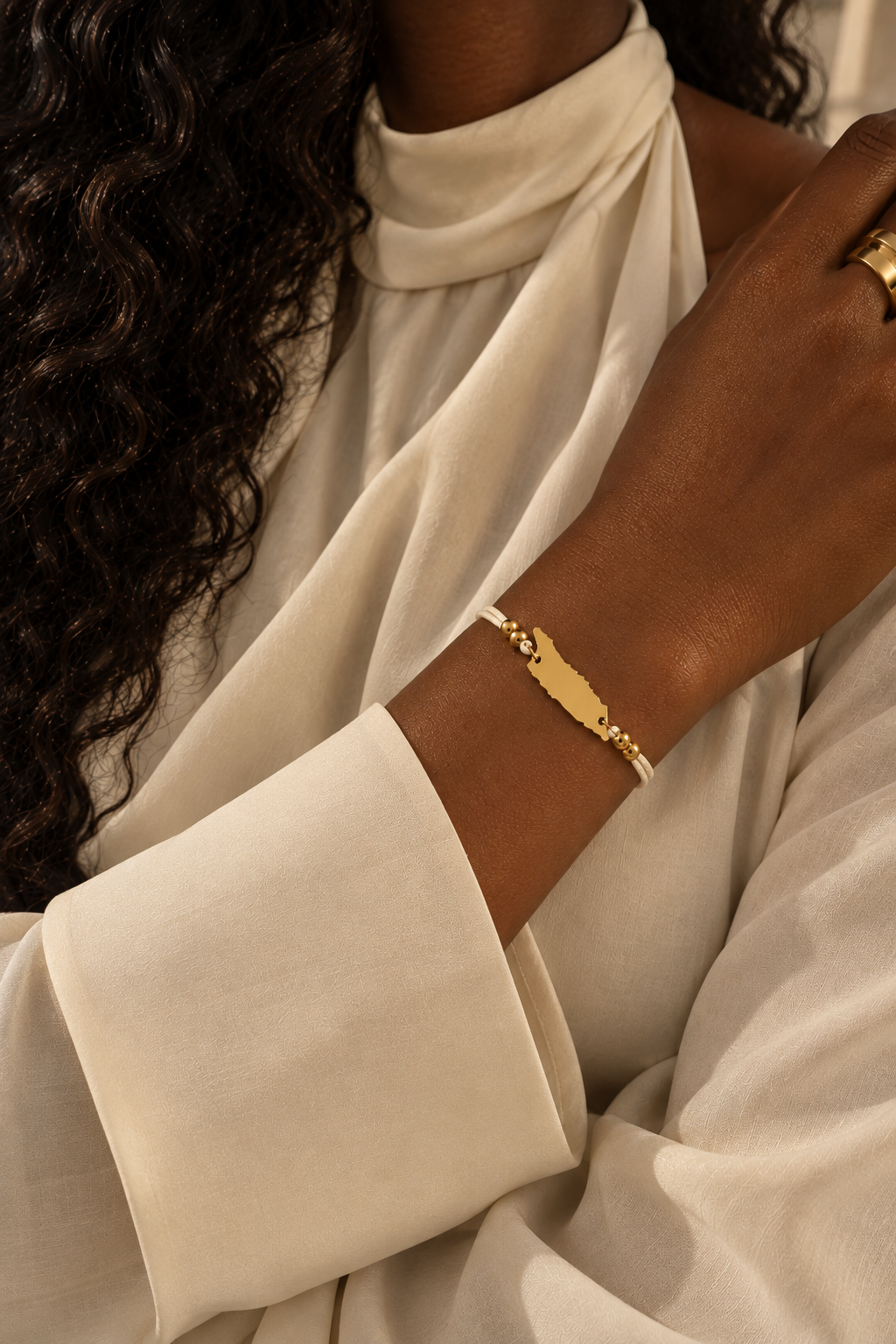 A close-up of a person wearing a gold-tone bracelet with map silhouette of Aruba  on the wrist, paired with a cream-colored draped blouse.