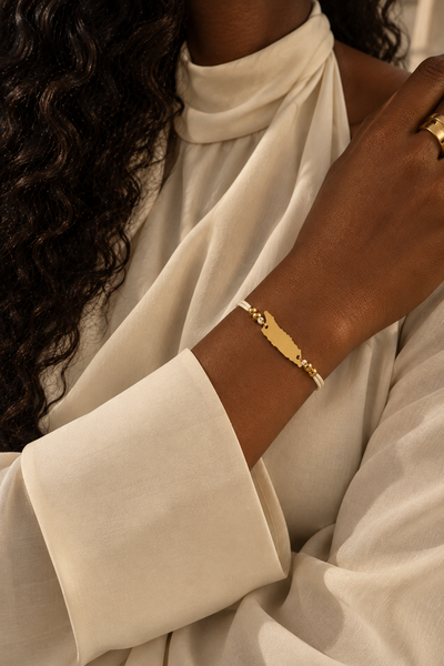 A close-up of a person wearing a gold-tone bracelet with map silhouette of Aruba  on the wrist, paired with a cream-colored draped blouse.