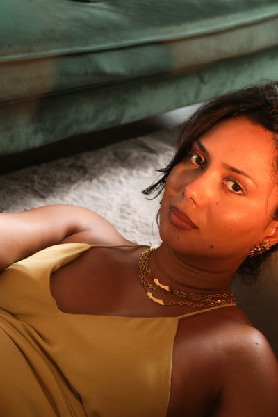 A woman wearing a mustard yellow satin dress and gold Concept Statement jewelry, lying on a gray rug in front of a green velvet sofa.