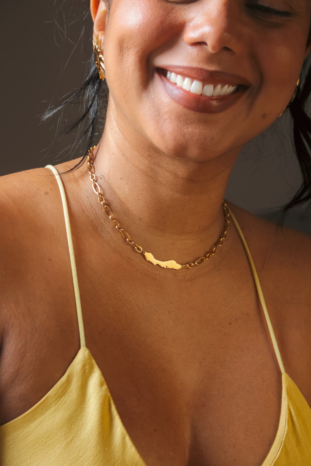 A close-up lifestyle shot of a woman wearing a delicate gold chain necklace with a small, polished gold Curacao island pendant by Concept Statement She is smiling and wearing a yellow camisole top against a dark, neutral background.