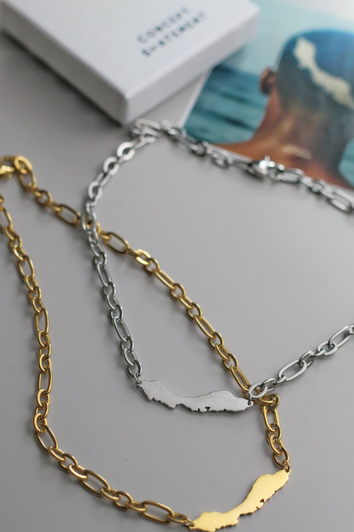 Two Curacao chain necklaces, one gold and one silver, on a gray surface with a blurred background.