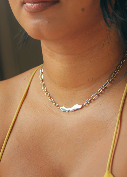 A close-up of a woman wearing a silver-toned chain necklace with a small, polished silver Curacao charm in the center, paired with a yellow bikini top.
