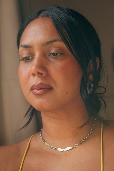 A close-up portrait of a woman wearing a Curacao silver chain necklace by Concept Statement with a small white wave-like pendant, styled with a yellow top against a soft beige background.