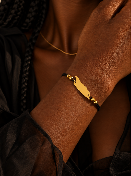 A close-up lifestyle shot of a model wearing a gold-plated map of Aruba bracelet on a black cord, paired with a delicate gold chain necklace.