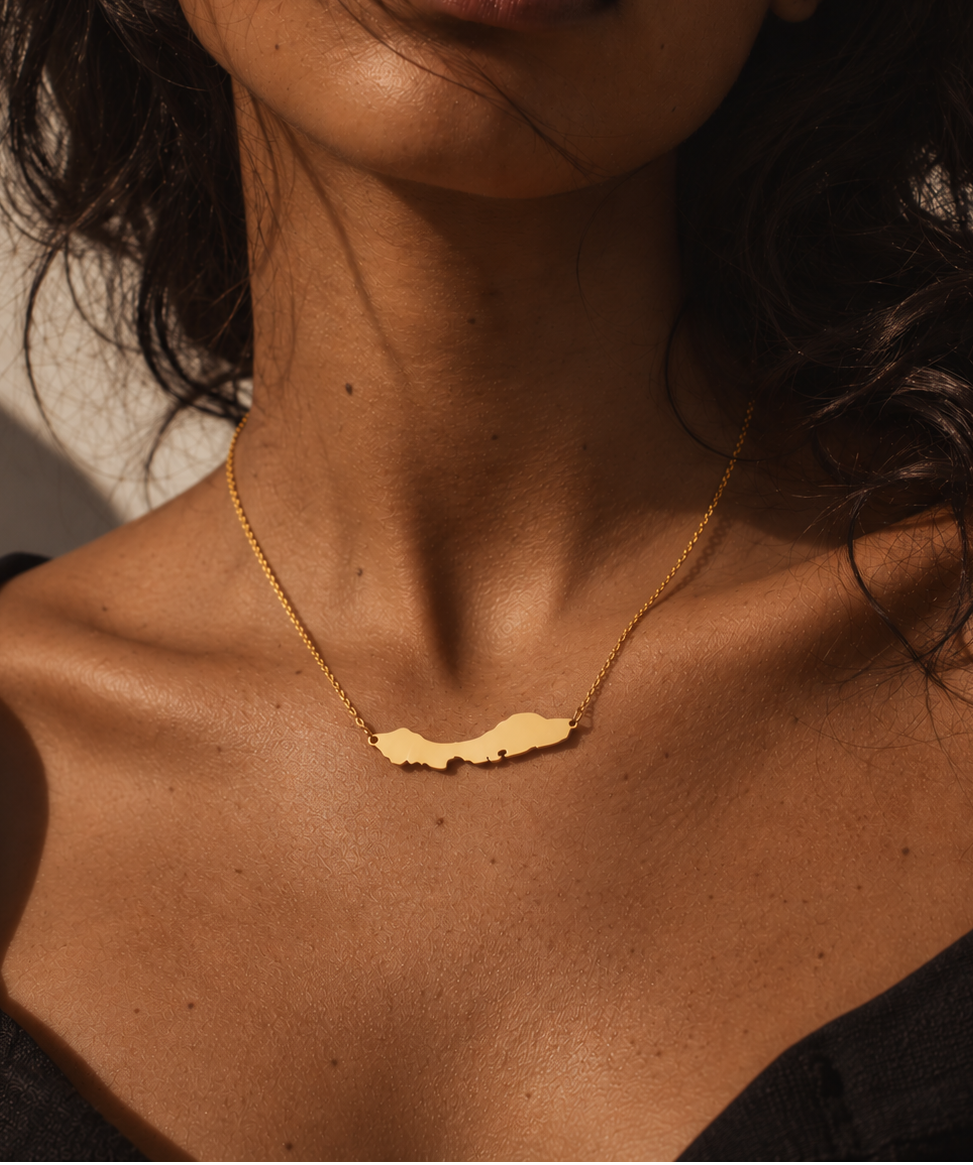 A close-up lifestyle shot of a model wearing a delicate gold chain necklace with a small, abstract Curacao pendant against warm sunlight.