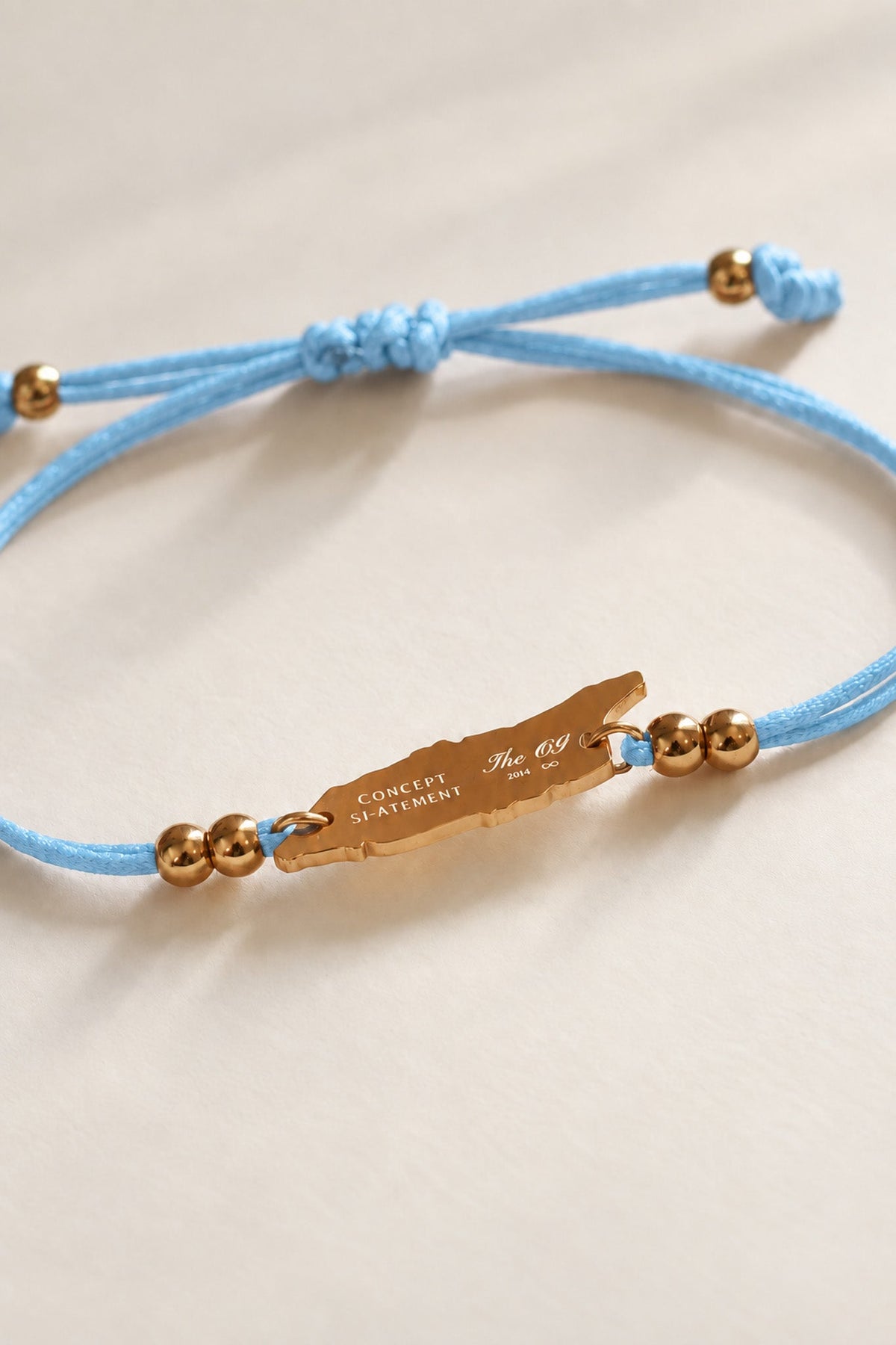 A light blue cord bracelet with a gold-toned engraved Concept Statement logo and OG map-shaped Aruba charm, gold beads, and a knotted sliding knot, shown on a neutral cream background.