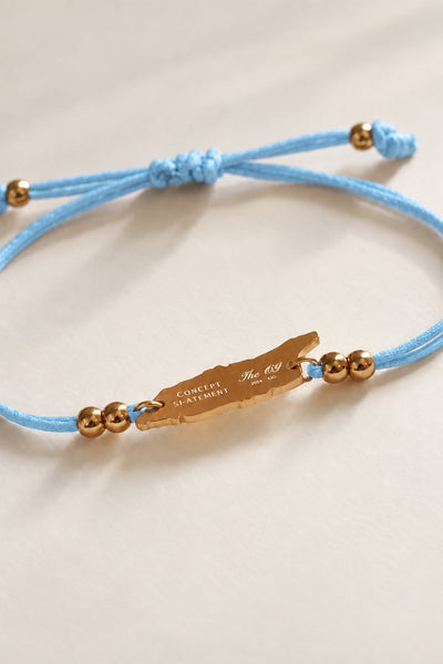 A light blue cord bracelet with a gold-toned engraved Concept Statement logo and OG map-shaped Aruba charm, gold beads, and a knotted sliding knot, shown on a neutral cream background.