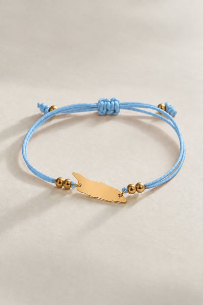 A light blue cord bracelet with a gold-tone Aruba charm and small gold beads, displayed on a neutral beige background.